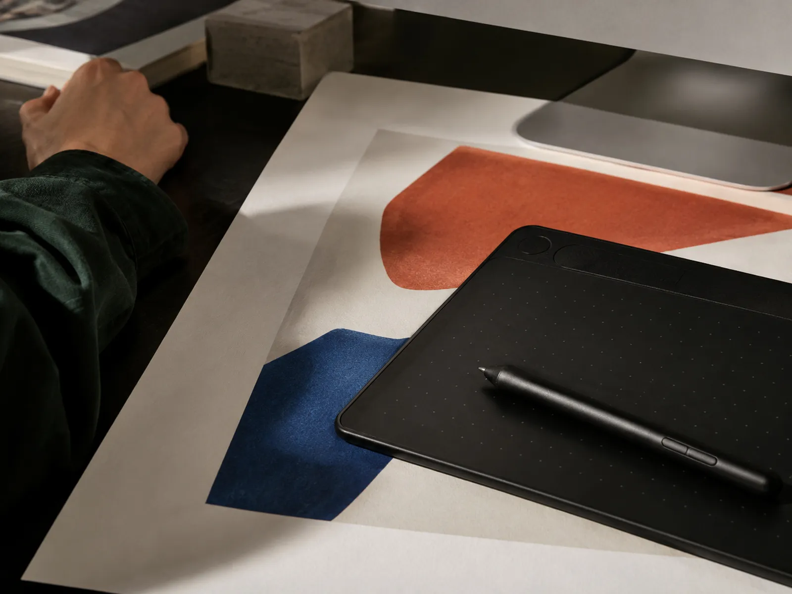 A drawing tablet and stylus on a desk beside printed artwork